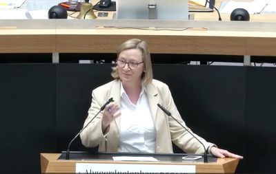 Eine Frau mittleren Alters mit blondem, schulterlangem Haar und Brille steht an einem Rednerpult im Abgeordnetenhaus Berlin. Sie trägt ein beiges Sakko über einer weißen Bluse und gestikuliert mit der linken Hand, während sie spricht. Auf dem Pult liegt ein Blatt Papier, daneben ein Smartphone. Hinter ihr sind Mikrofone, Unterlagen und ein großer Monitor zu sehen.