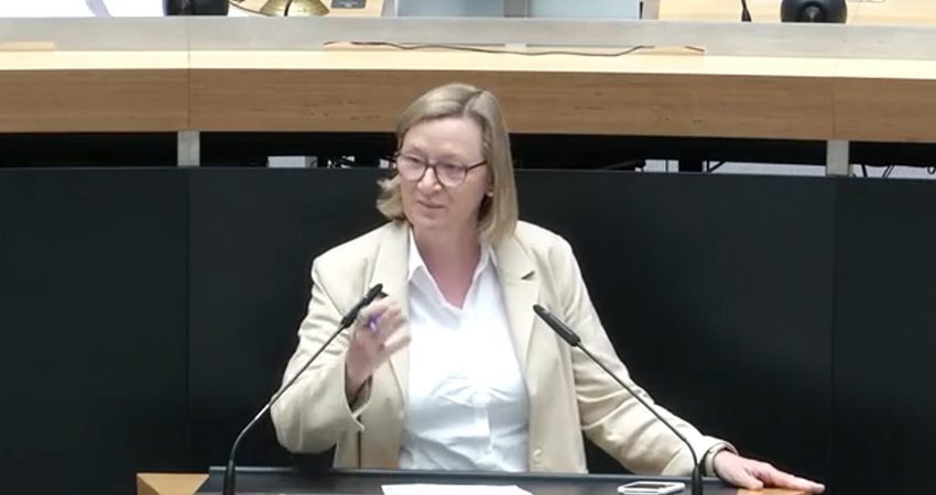 Eine Frau mittleren Alters mit blondem, schulterlangem Haar und Brille steht an einem Rednerpult im Abgeordnetenhaus Berlin. Sie trägt ein beiges Sakko über einer weißen Bluse und gestikuliert mit der linken Hand, während sie spricht. Auf dem Pult liegt ein Blatt Papier, daneben ein Smartphone. Hinter ihr sind Mikrofone, Unterlagen und ein großer Monitor zu sehen.
