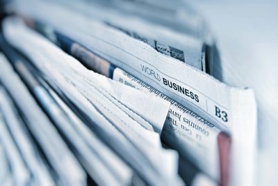 Ein Stapel eng gefalteter Tageszeitungen, bei dem eine Ausgabe mit der sichtbaren Überschrift ‚WORLD BUSINESS‘ und der Seitenangabe ‚B3‘ im Fokus steht. Die Farben sind überwiegend in Blautönen gehalten, was dem Bild eine kühle, sachliche Atmosphäre verleiht.