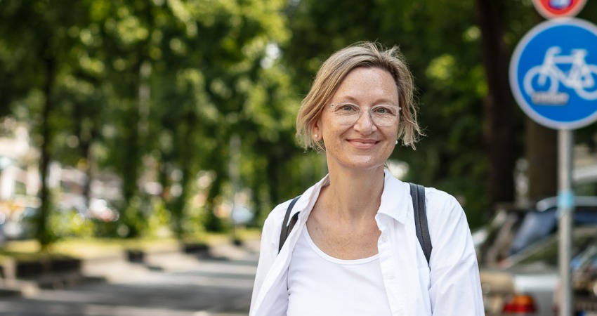 Petra Vandrey steht vor grünen Bäumen. Ein Fahrradschild ist auf der Klinkenseite zu sehen.