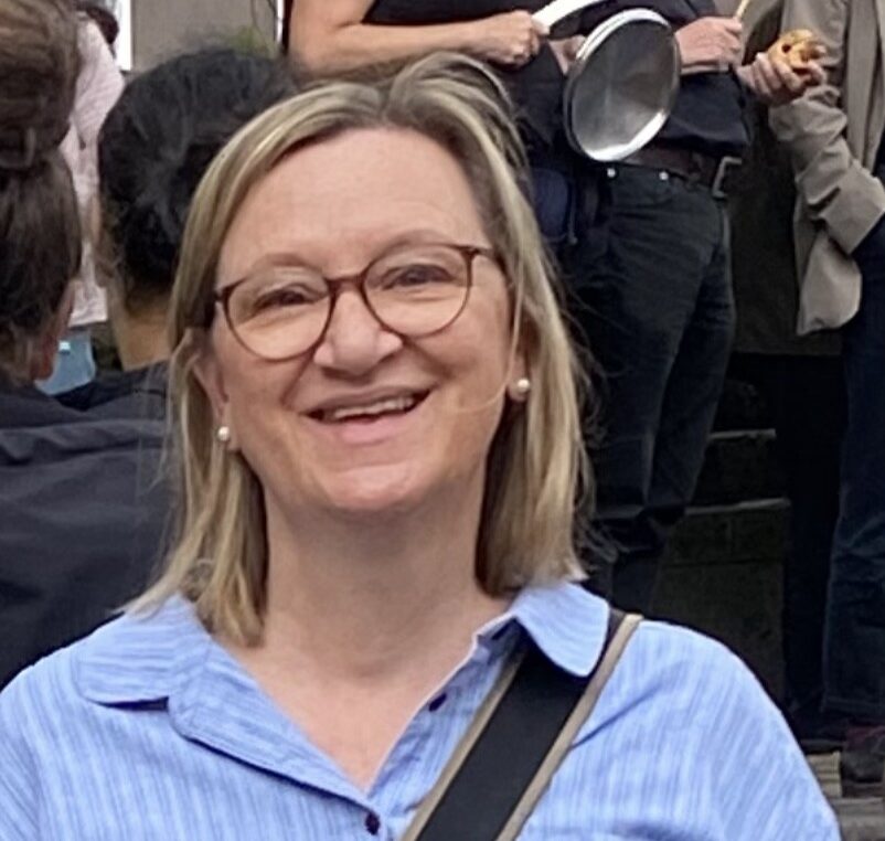 Eine lächelnde Frau mit blonden, schulterlangen Haaren, Brille und blauer Bluse steht im Freien vor einer Gruppe von Menschen.